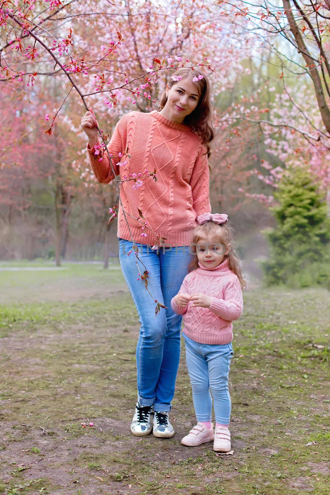 Happy mother and little daughter stand in park under pink cherry blossom . Vertical