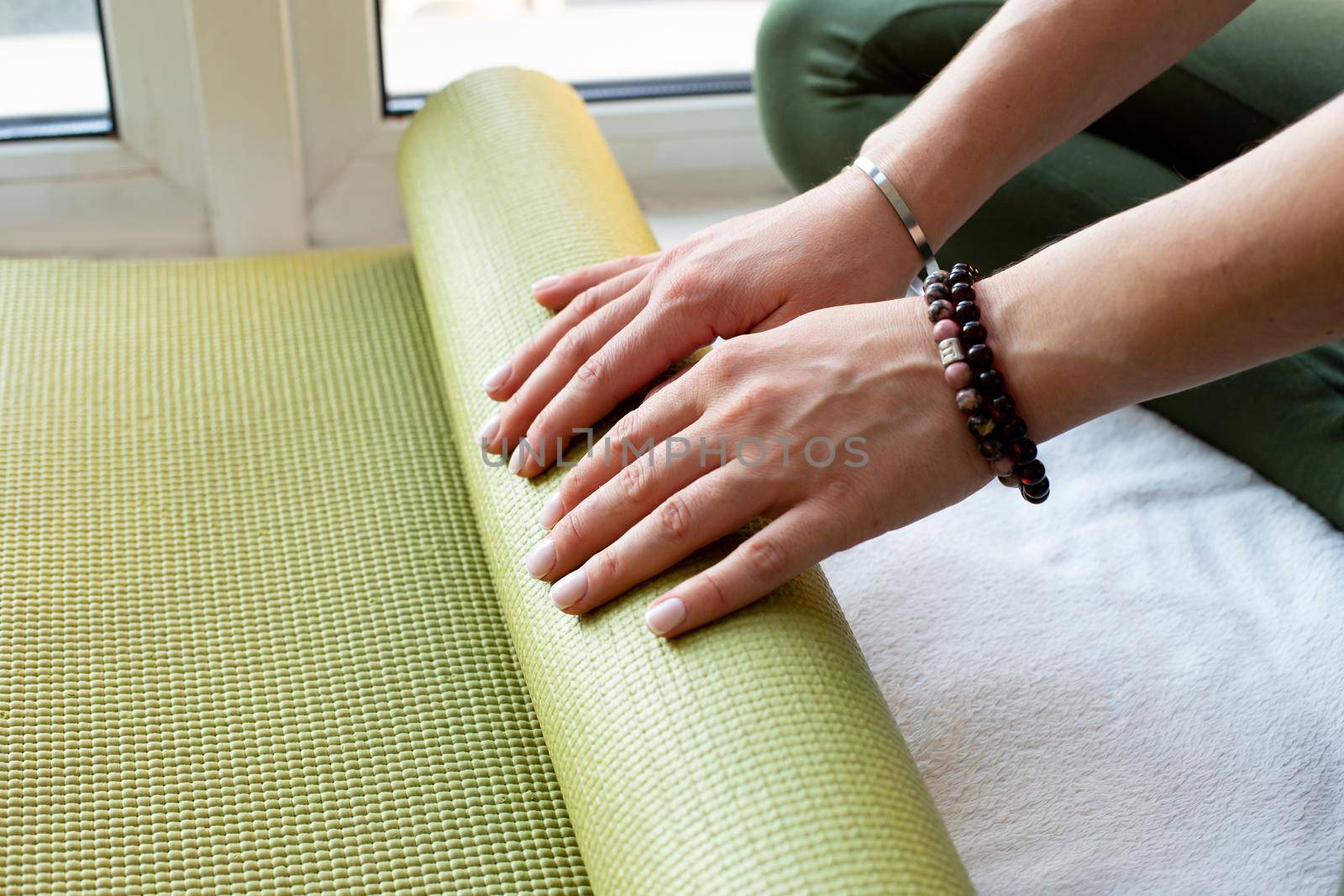 A hands slender woman twirls green rubber mat for fitness or yoga sitting in a bright room by the window. Close up