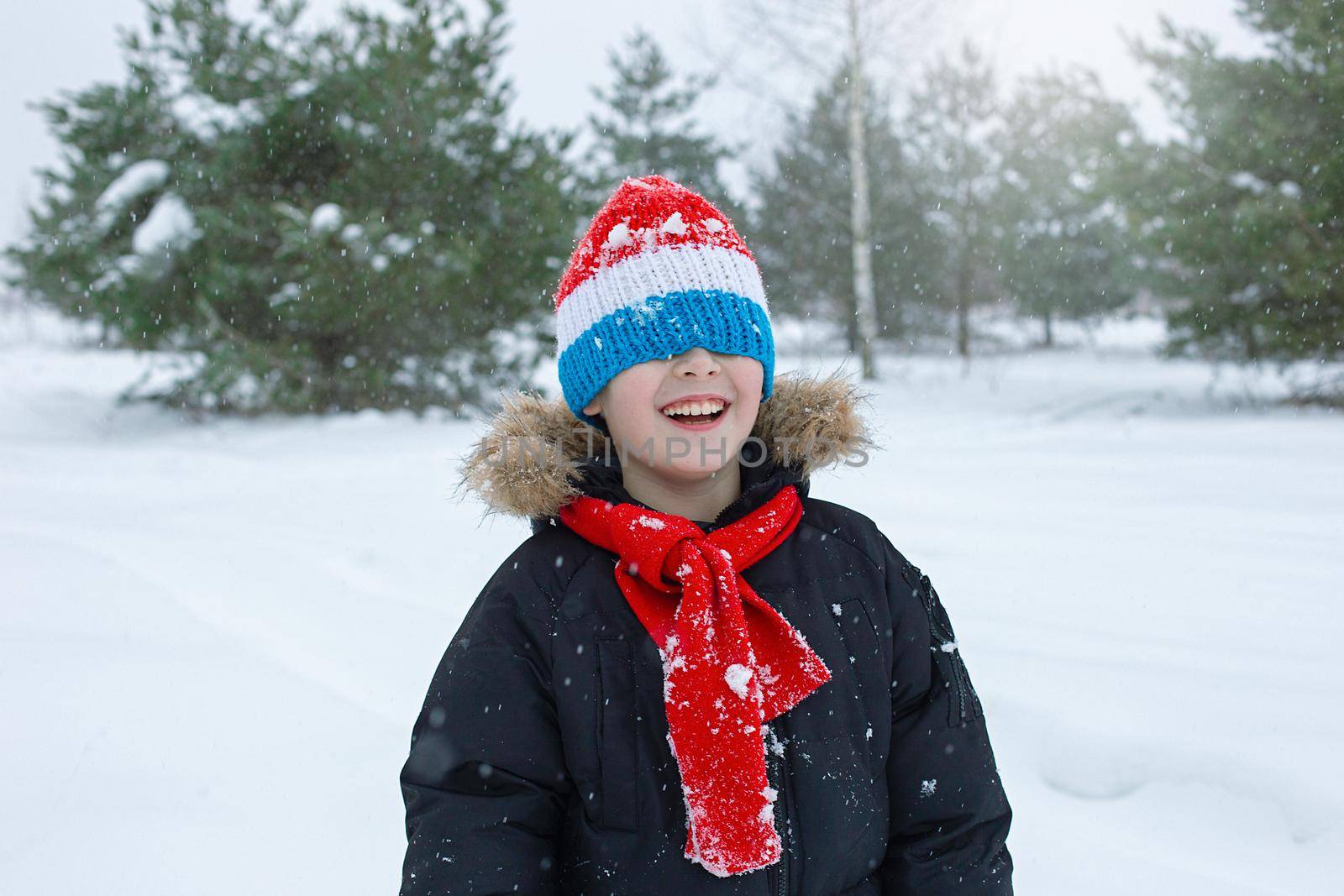 Portrait of a happy boy in bright winter clothes and a red scarf stands in a striped hat pulled down over his eyes, smiling, in a winter park with pine trees