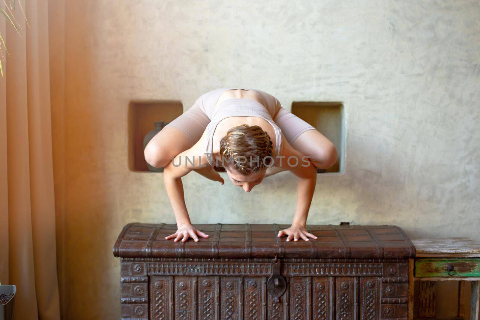 A beautiful and slender woman in a beige top and breeches, stands in a yoga pose, performs a handstand exercise, in a room with an oriental interior