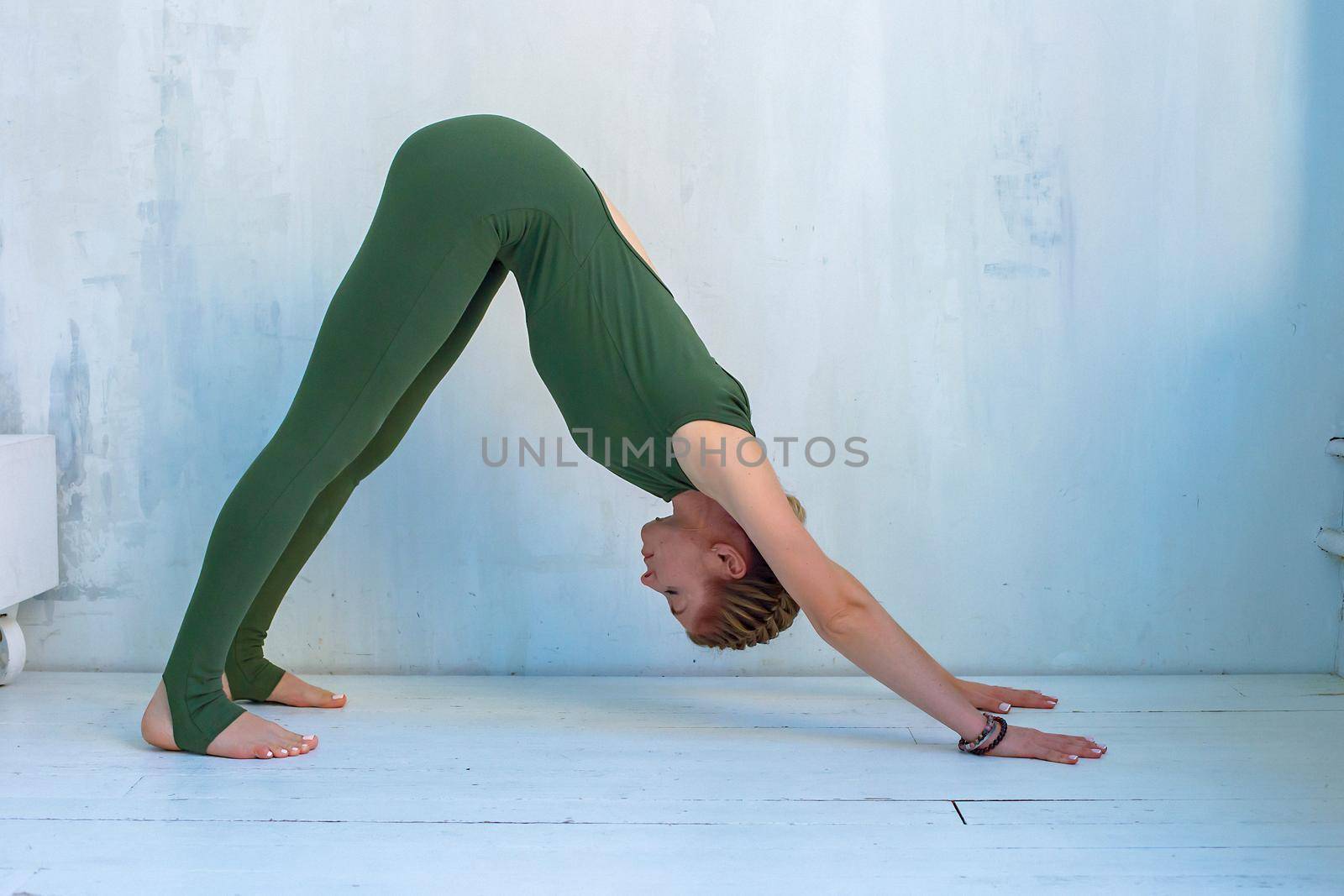 A beautiful and slender girl in green sportswear performs a pose in yoga -dog face down, near a light blue wall