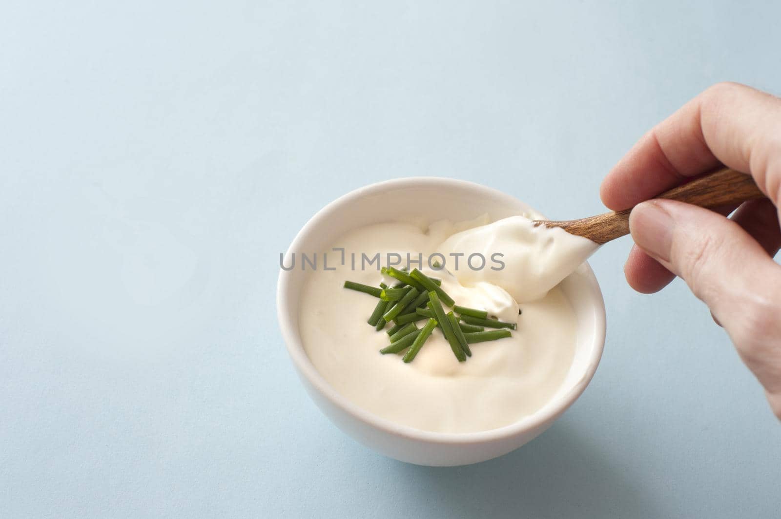 Man dipping a small spoon into a white ceramic bowl of sour cream garnished with fresh chopped chives over a grey background with copy space