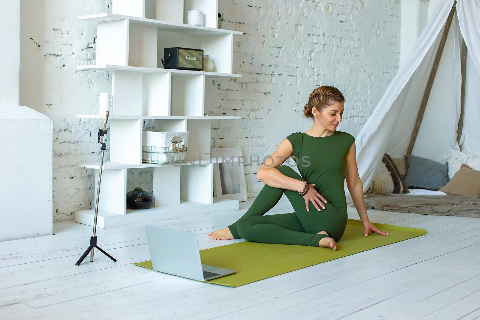 A slender woman in a green sports jumpsuit performs a yoga exercise at home, in a light interior, in front of a laptop. Conducts online classes using a smartphone