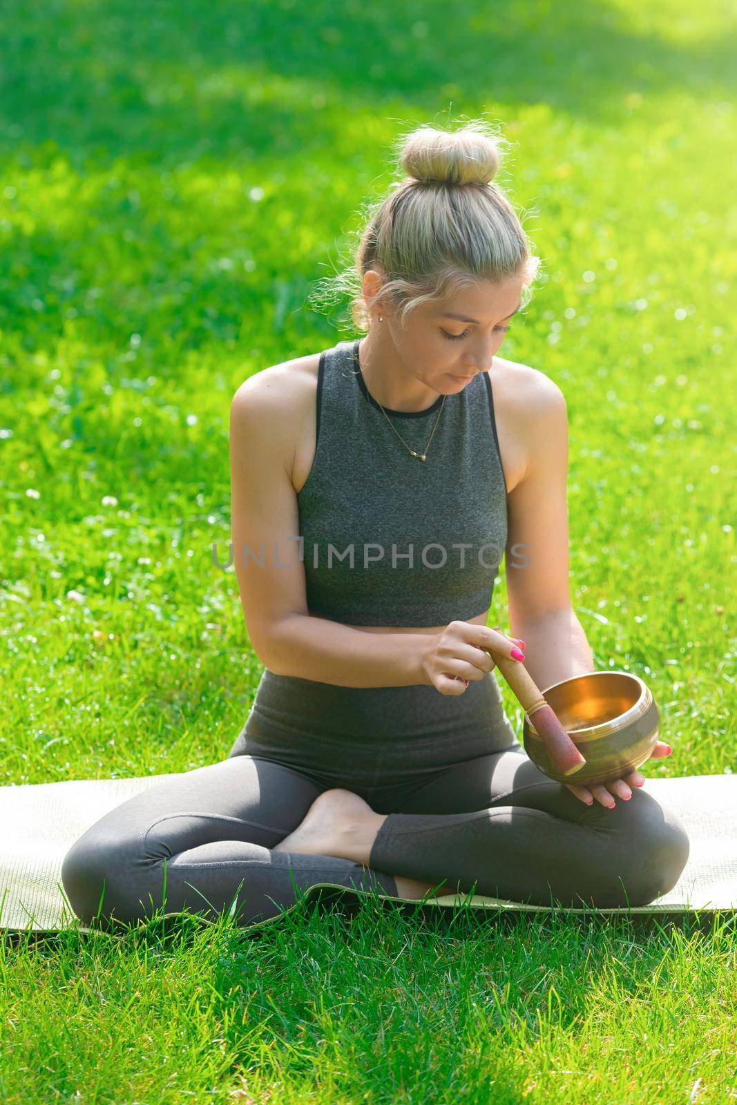 A blonde woman in a gray top and leggings, sitting on the green grass in summer, holds a meditation bowl. Vertical