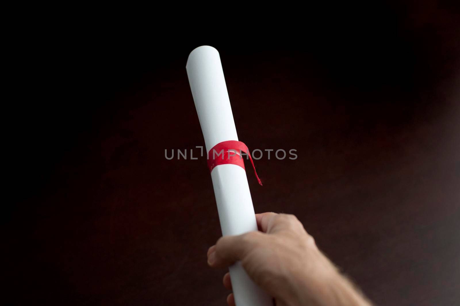 Studio shot over black of a a male hand holding a diploma or document, rolled and tied with red ribbon