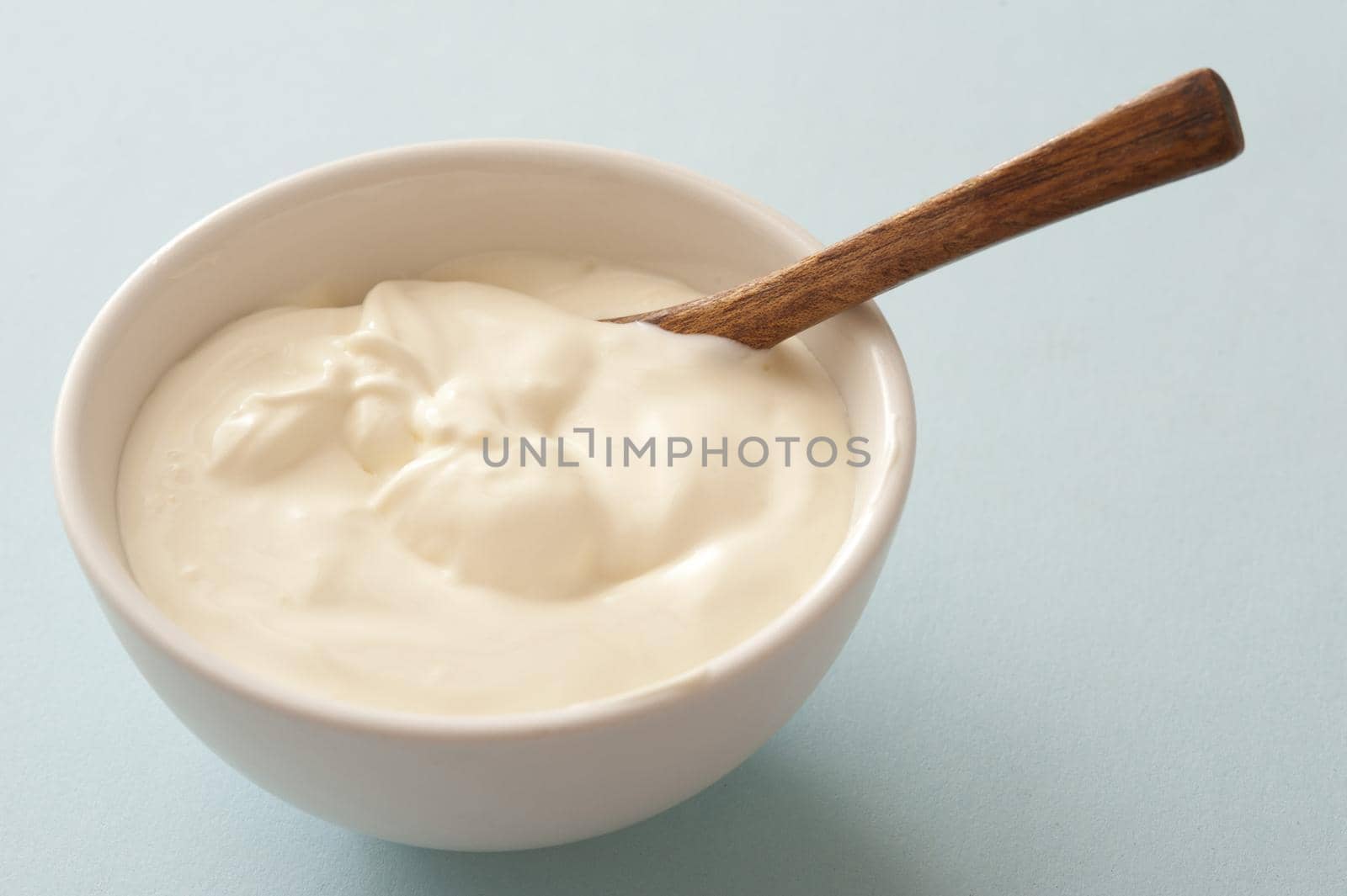 Plain white ceramic bowl of sour cream with a spoon over a grey background for use as a topping and ingredient in cooking