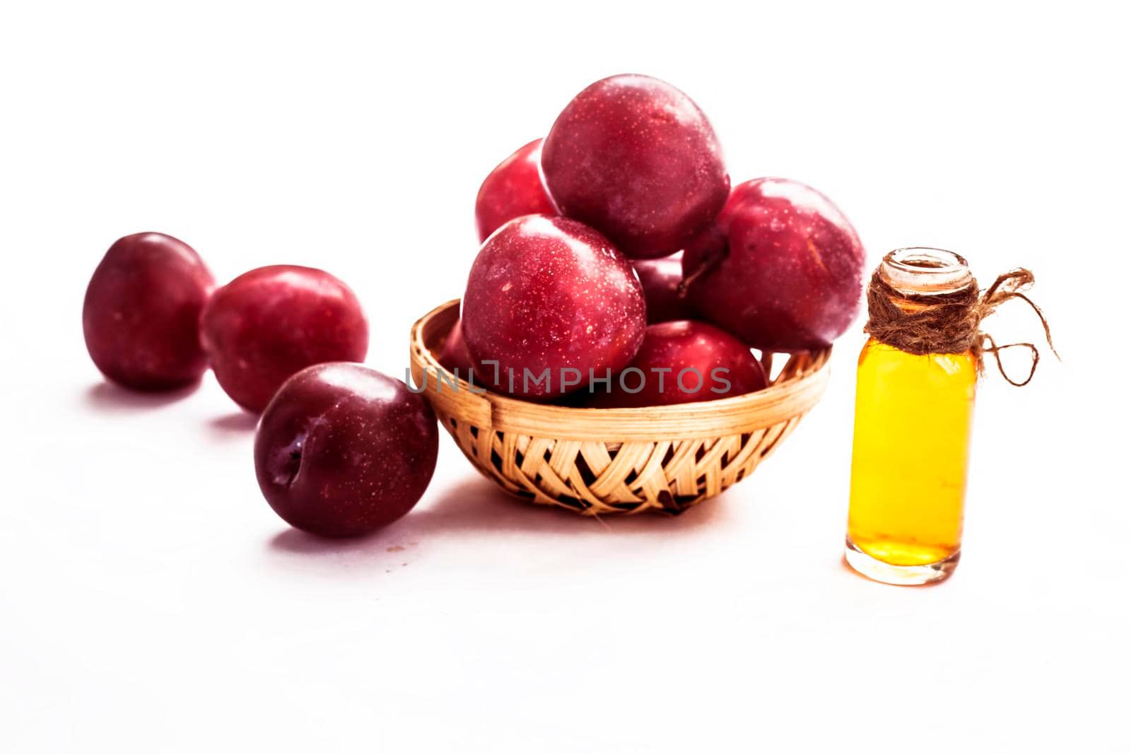 Raw organic red ripe plums in a brown-colored basket along with its extracted essential oil in a transparent glass bottle isolated on white.