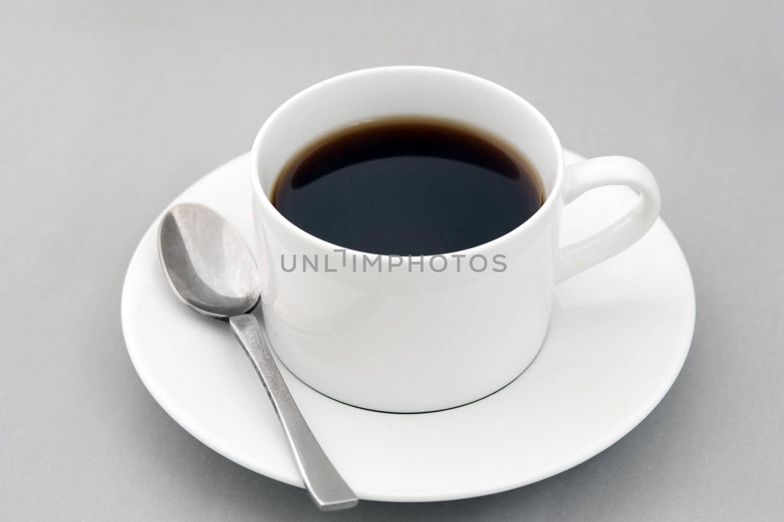 Cup of strong black espresso coffee served in a generic white ceramic cup and saucer with teaspoon over a grey background