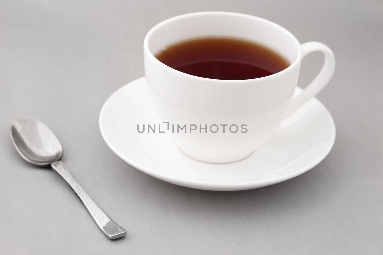 Generic white cup and saucer of hot black tea with a teaspoon alongside over gray