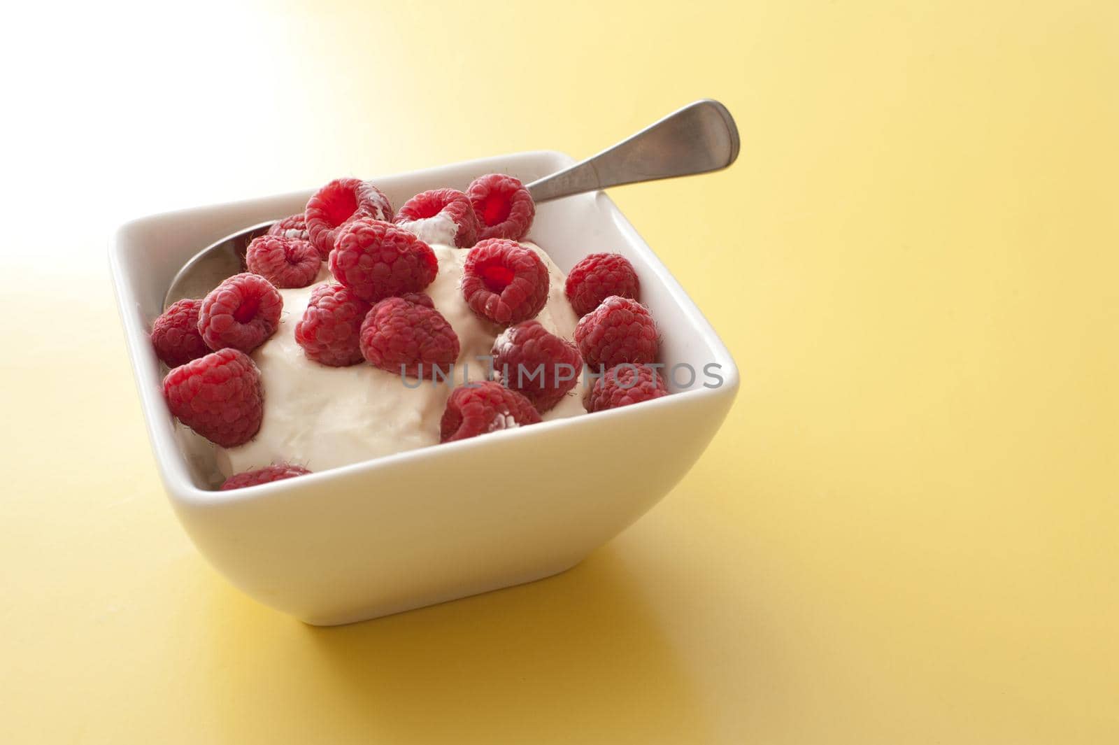 Delicious creamy yoghurt topped with fresh red raspberries served in a modern square bowl over a yellow background with copy space
