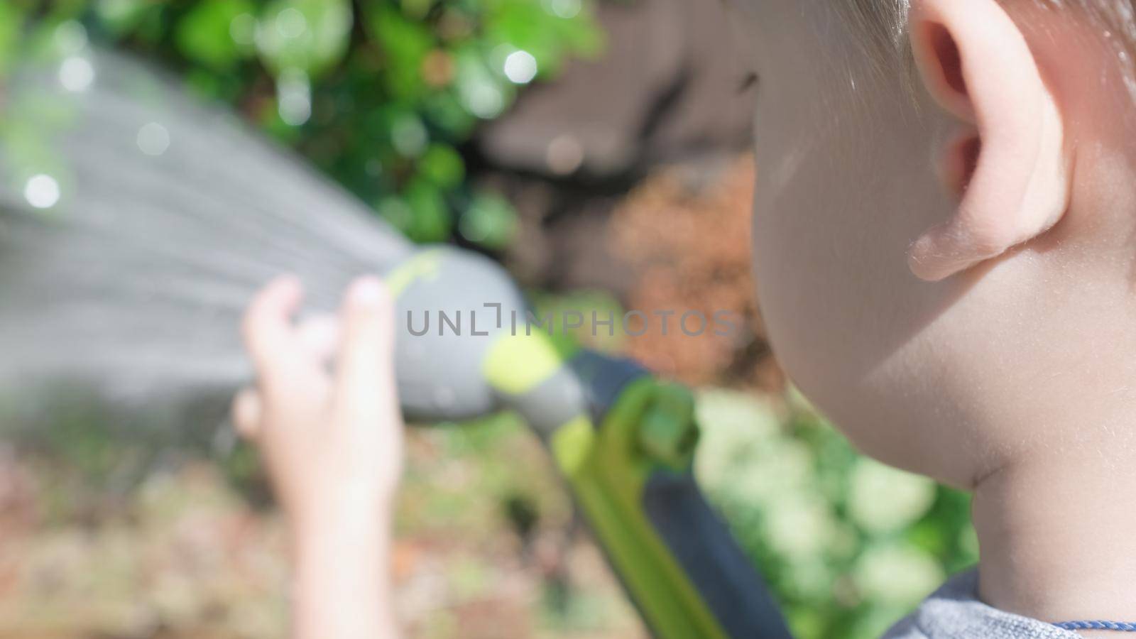 Funny little boy watering lawn plants in garden housing backyard. Adorable child playing with irrigation hose at hot sunny summer outdoors. Children help with housework. activity for kids. Childhood.