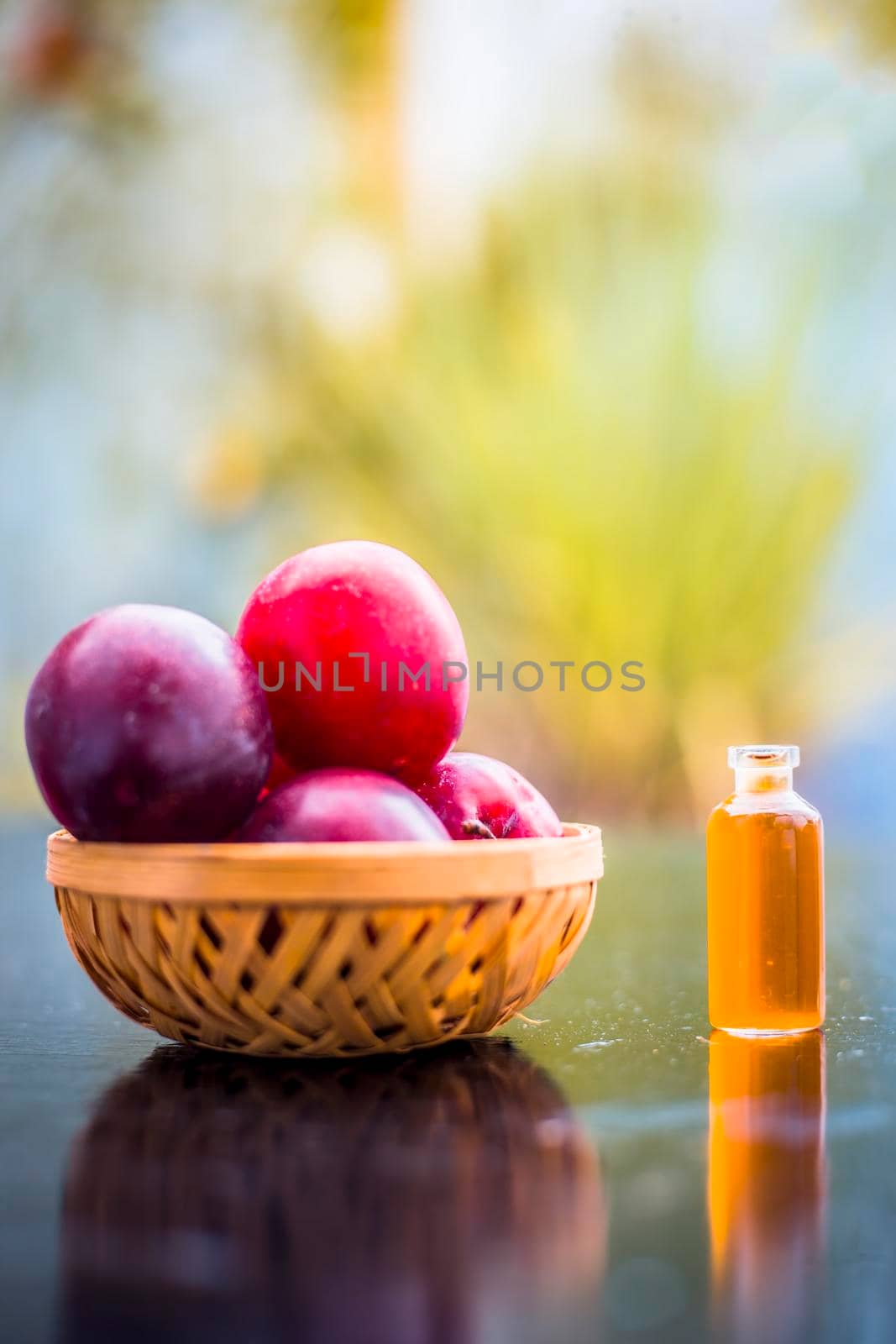 Raw organic red ripe plums in a brown-colored basket on the wooden surface along with its extracted essential oil in a small transparent bottle with blurred background.