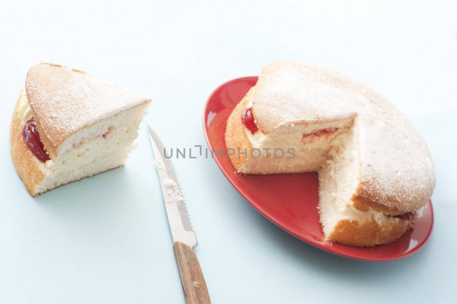 Freshly baked jam sponge cream cake with a generous slice on the side alongside a knife, served on a red plate
