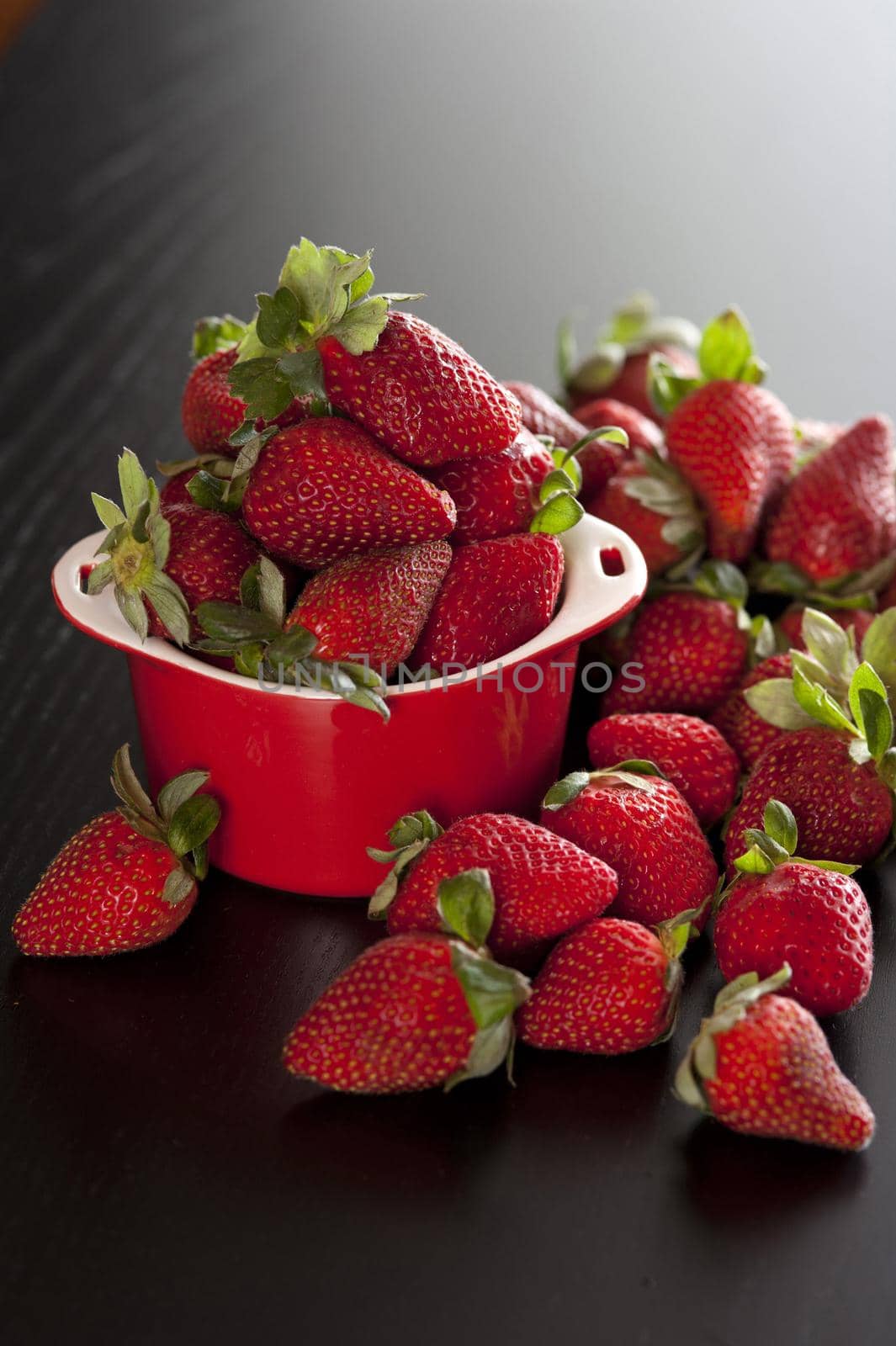 Ripe red strawberries in a small ramekin overflowing onto a dark surface with a highlight behind for copyspace