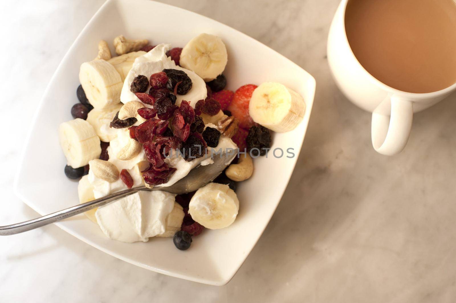 Healthy breakfast serving of assorted fruit with blueberries, strawberries , sliced fresh banana and raisins on fresh creamy yoghurt with tea