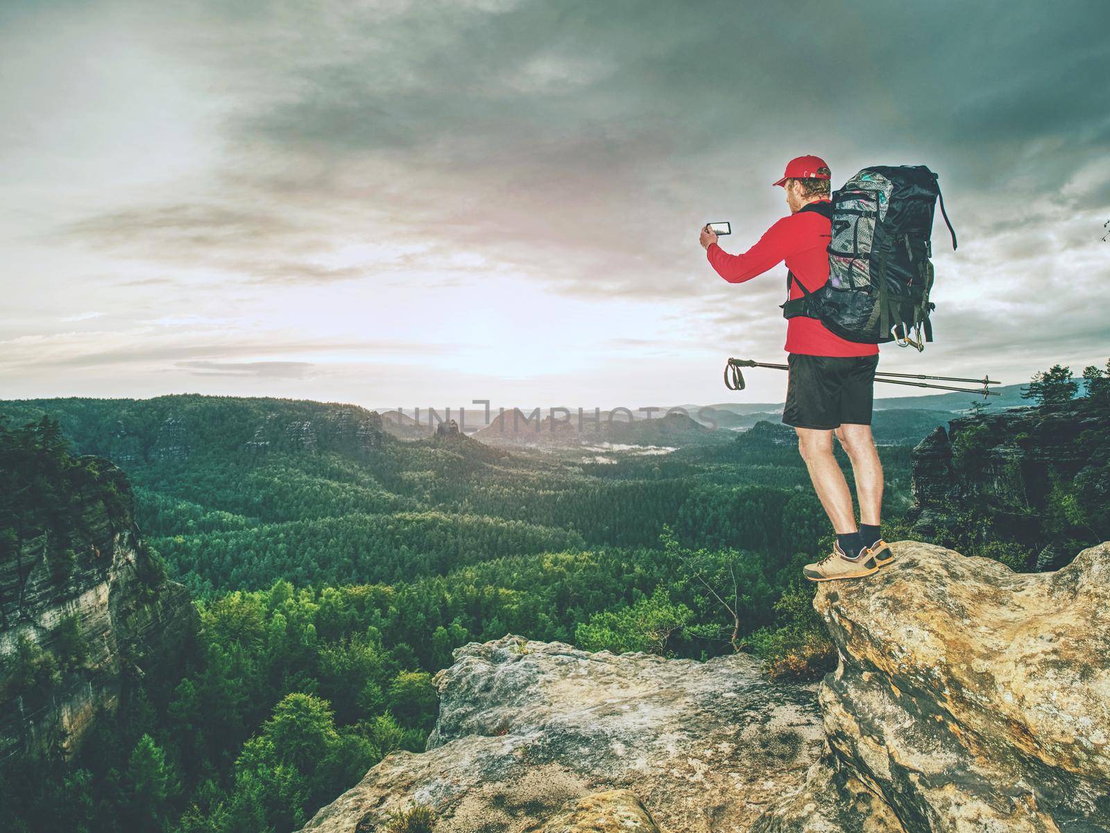 Hiker is taking photo by smart phone on the peak of mountain. Man in red t-shirt and shorts takes photos with smart phone on peak of rock empire. Dreamy fogy landscape 