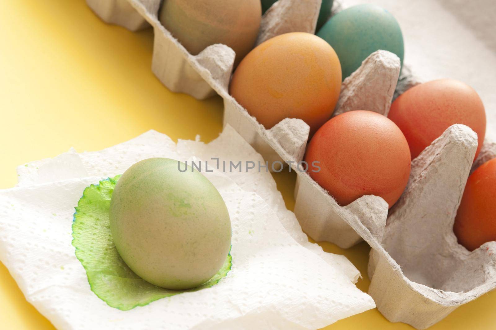 Dyeing colorful homemade boiled Easter eggs in a country tradition with a cardboard box full alongside a single green egg drying on a paper napkin