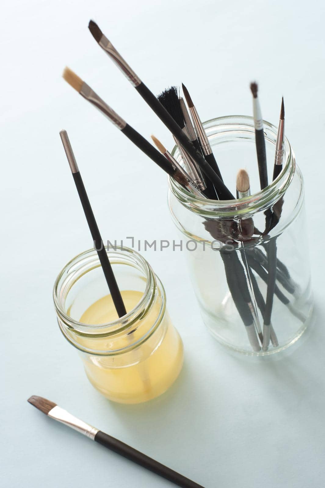 Top down view of various sized artist paintbrushes in jars and thinner solution over white table