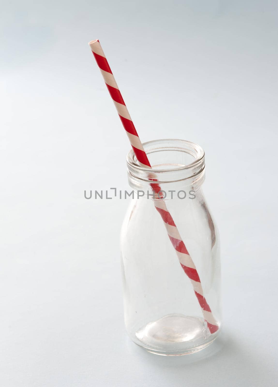 Empty kids glass milk bottle with striped red and white plastic straw over a white background with copy space