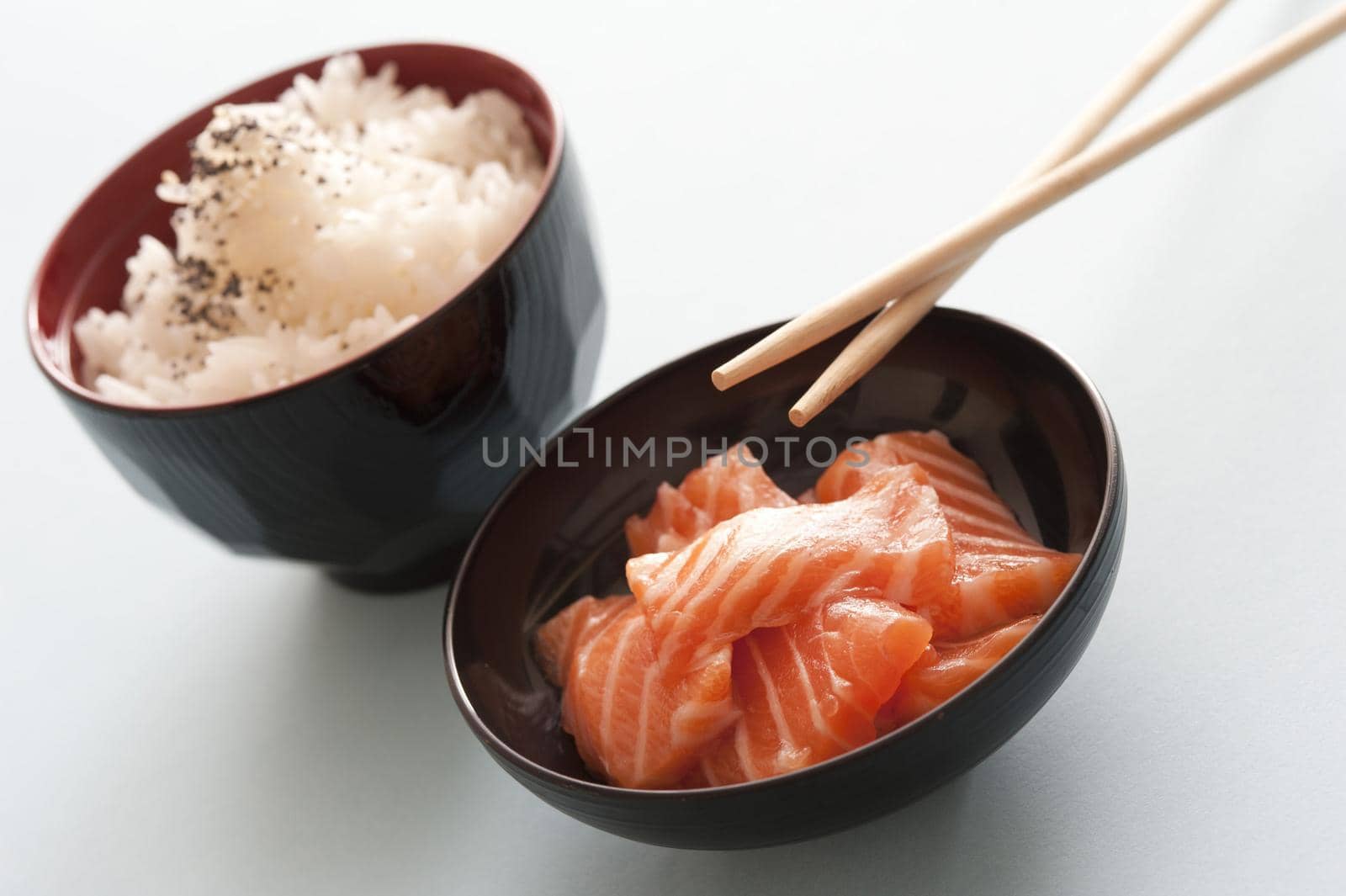 One black bowl of cubed pink salmon and another of steamed rice beside chopsticks against a white background