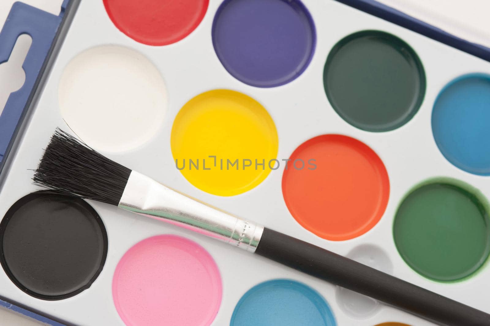 Box of new kids watercolor paints with colorful round containers of paint and an unused paintbrush view from above