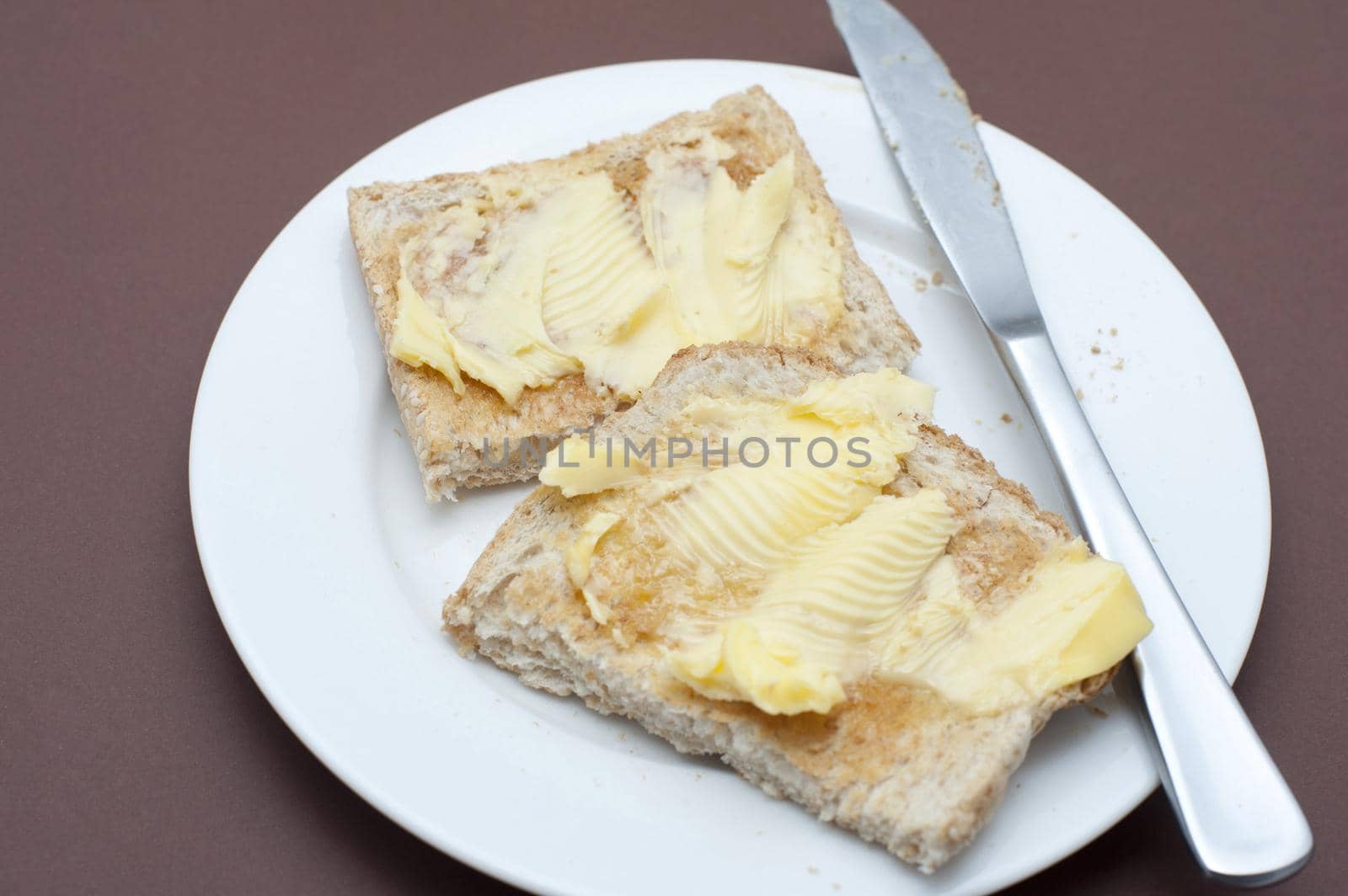 Close up Tasty Toasted Bread with Butter on Top Served on White Round Plate with Knife. Isolated on Brown Background.