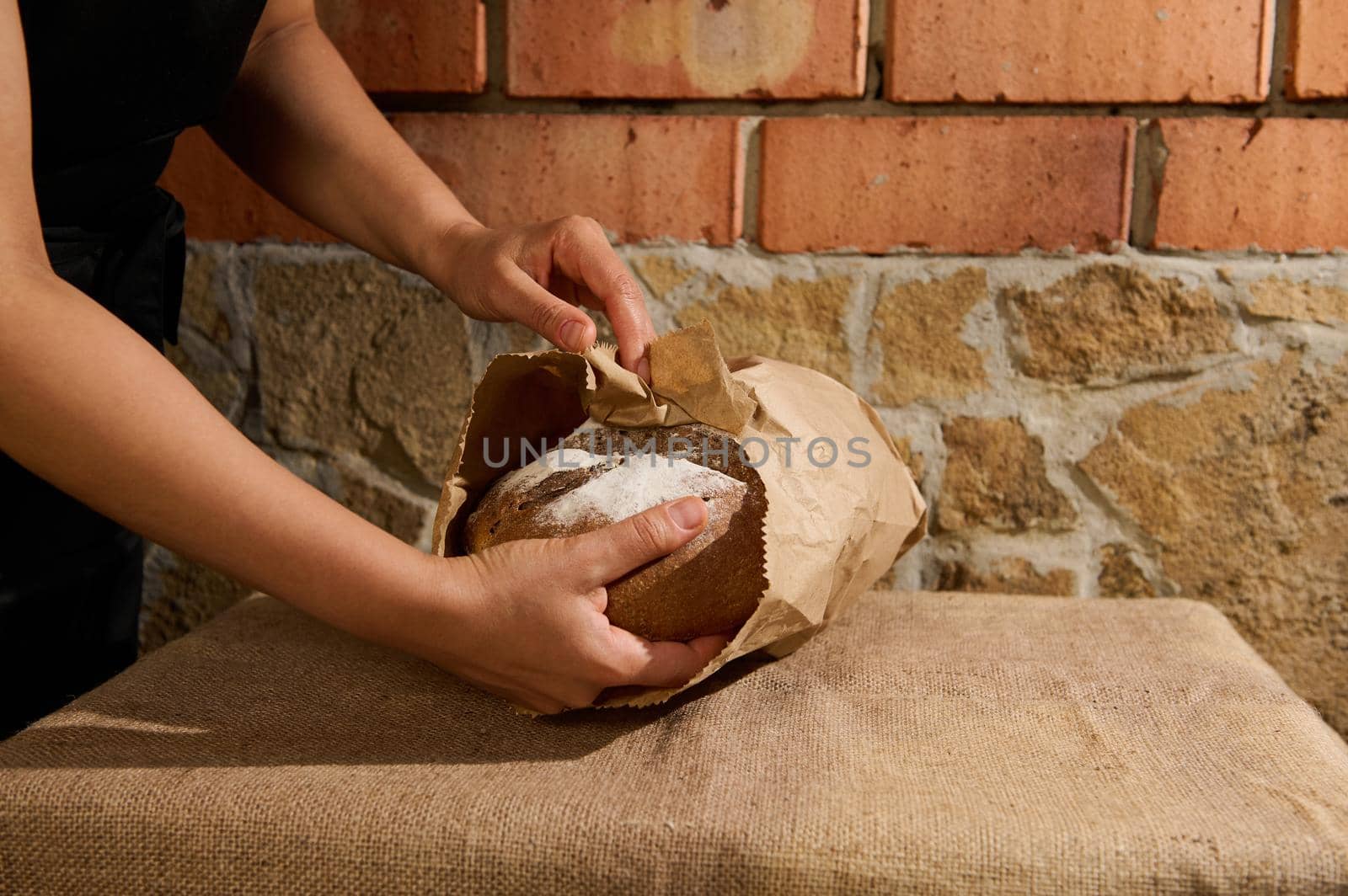 Selective focus. Close-up of hands of a baker confectioner putting healthy fresh homemade whole grain wheat bread into a recyclable eco paper bag, standing against brick and stone wall background