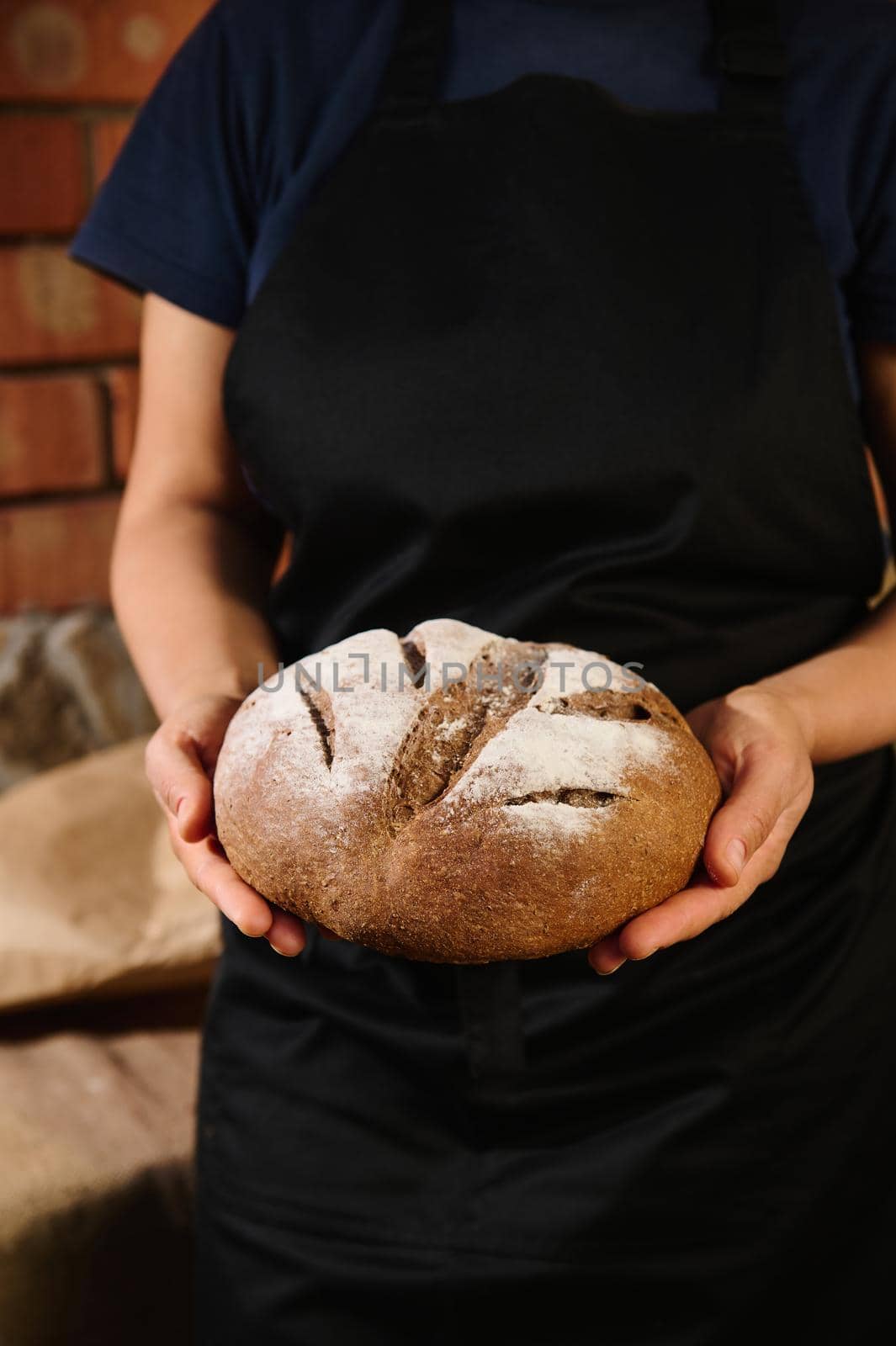 Close-up. Selective focus on an artisan loaf of traditional homemade sourdough boule rye bread with crust homemade rye bread in the hands of a woman baker dressed in black chef's apron.