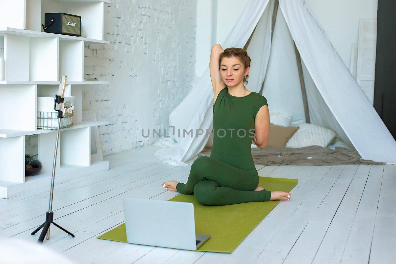 A slender woman in a green sports jumpsuit performs a yoga exercise at home, in a light interior, in front of a laptop. Conducts online classes using a smartphone
