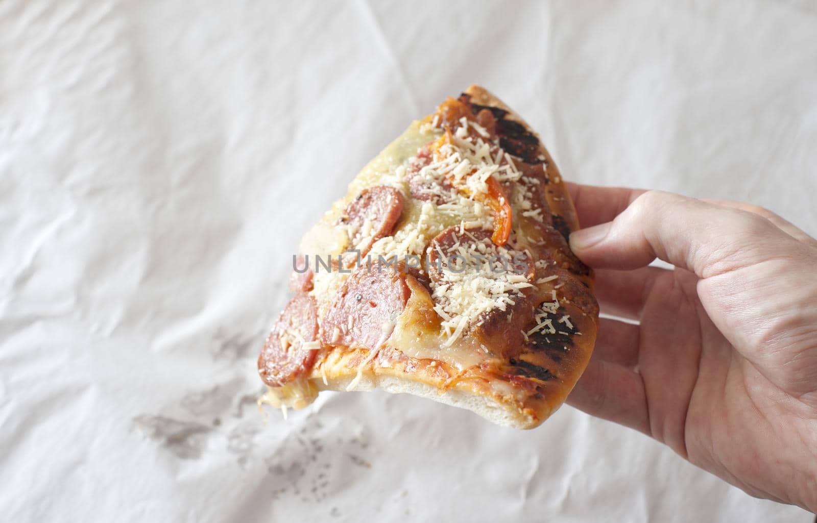 Man holding the last slice of salami or pepperoni pizza topped with spicy sausage, cheese and onion over crumpled white paper