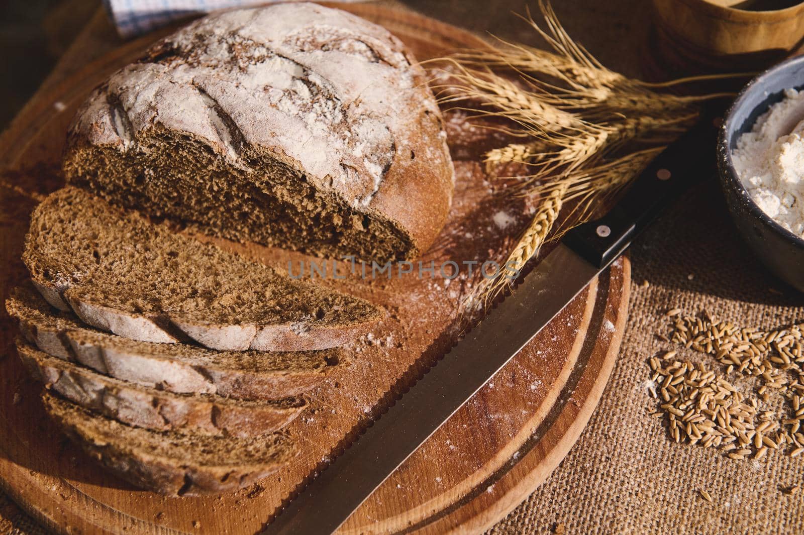 Still life with sliced loaf of a homemade whole grain sourdough rye bread, a kitchen knife on a wooden cutting board, and wheat ears near a bowl with white flour and scattered cereal grains on burlap