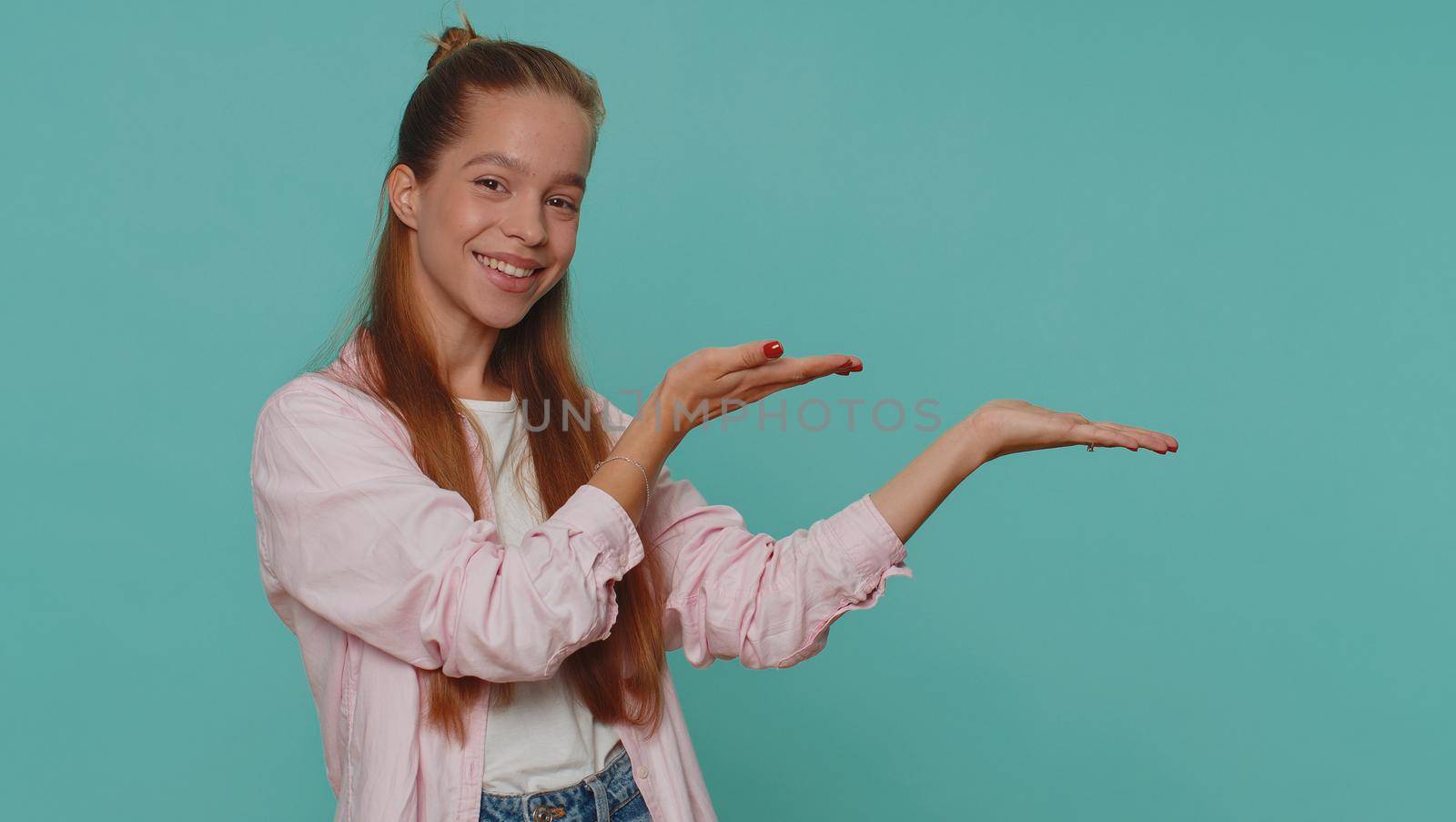 Lovely pretty teenager girl showing thumbs up and pointing right empty place, advertising area for commercial text, copy space for goods promotion. Young child kid isolated on blue studio background