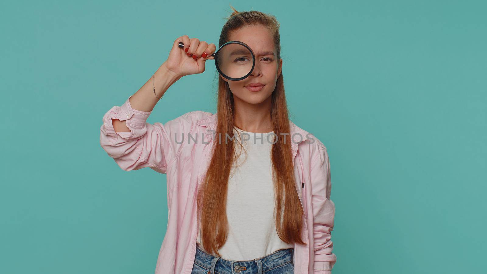 Investigator researcher scientist girl holding magnifying glass near face, looking into camera with big zoomed funny eye, searching, analysing. Young child kid on blue studio wall background indoors