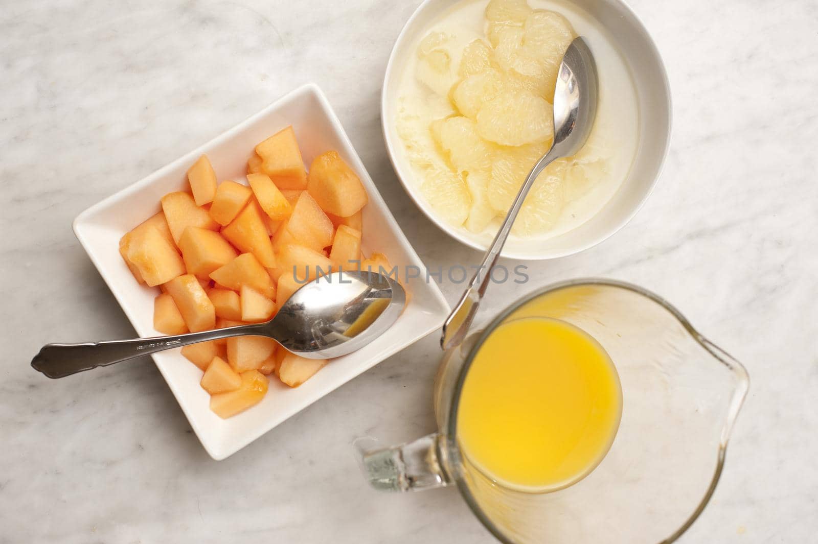 Variety of breakfast fruits with orange juice in a jug, and fresh diced melon and pears in individual dishes, overhead view
