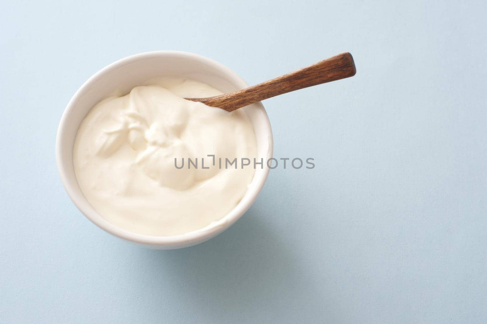 Small white round bowlk of plain yogurt with wooden spoon against a light colored background