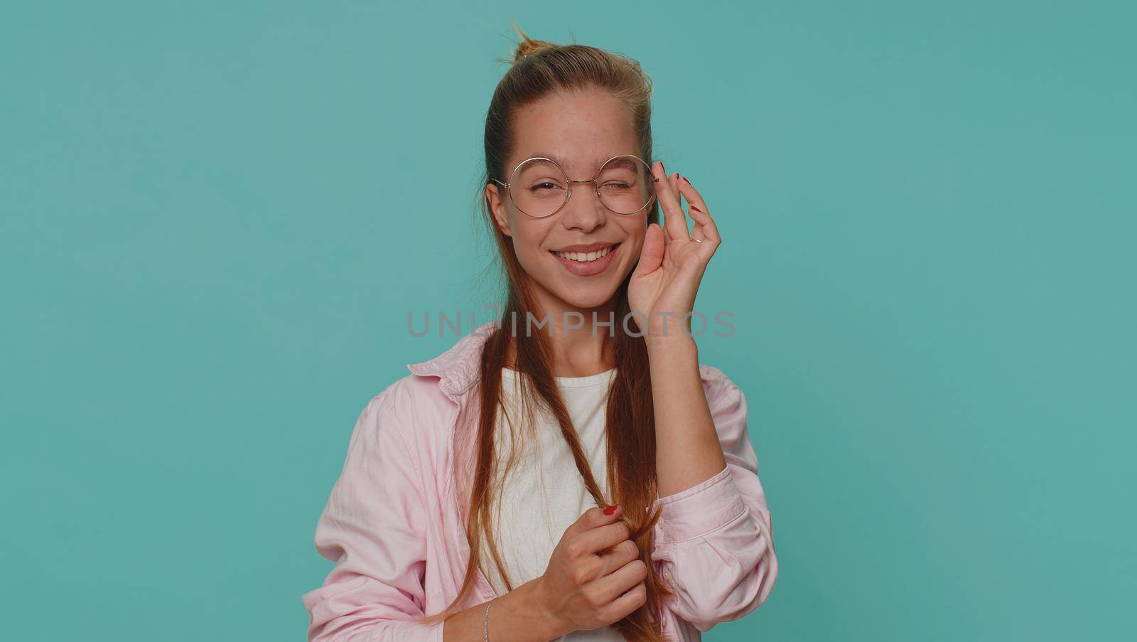 Smiling playful happy girl blinking eye, looking at camera with toothy smile, winking and flirting, expressing optimism. Young stylish female child kid isolated alone on blue studio background indoors