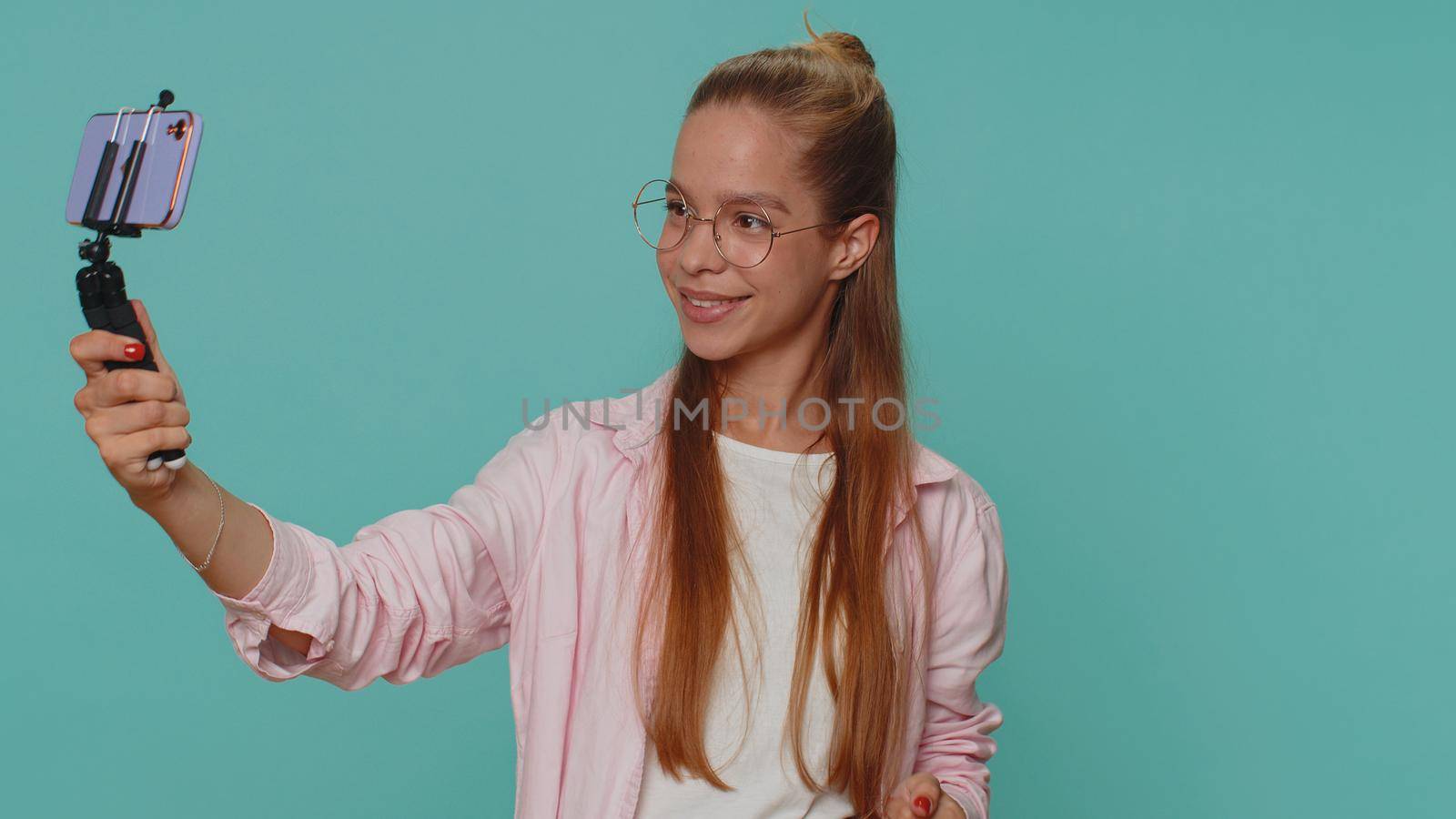 Pretty teenager girl traveler blogger taking selfie on mobile phone selfie stick, communicating video call online with subscribers. Young child kid isolated alone on blue studio background indoors