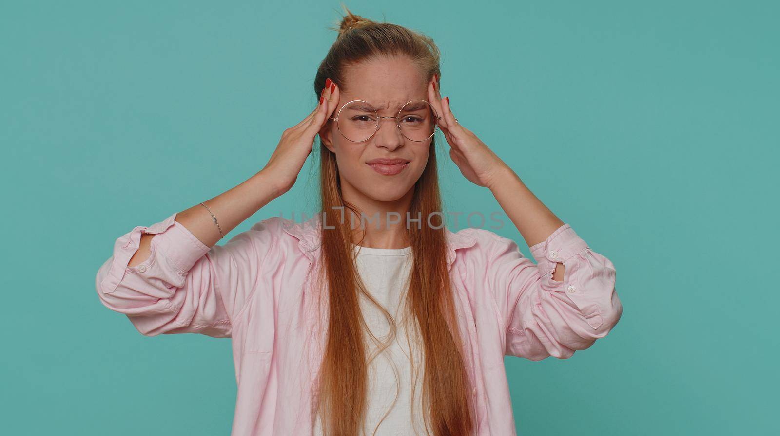 Displeased teenager young girl rubbing temples to cure headache problem, suffering from tension migraine, stress, grimacing in pain, high blood pressure. Student child kid on blue studio background