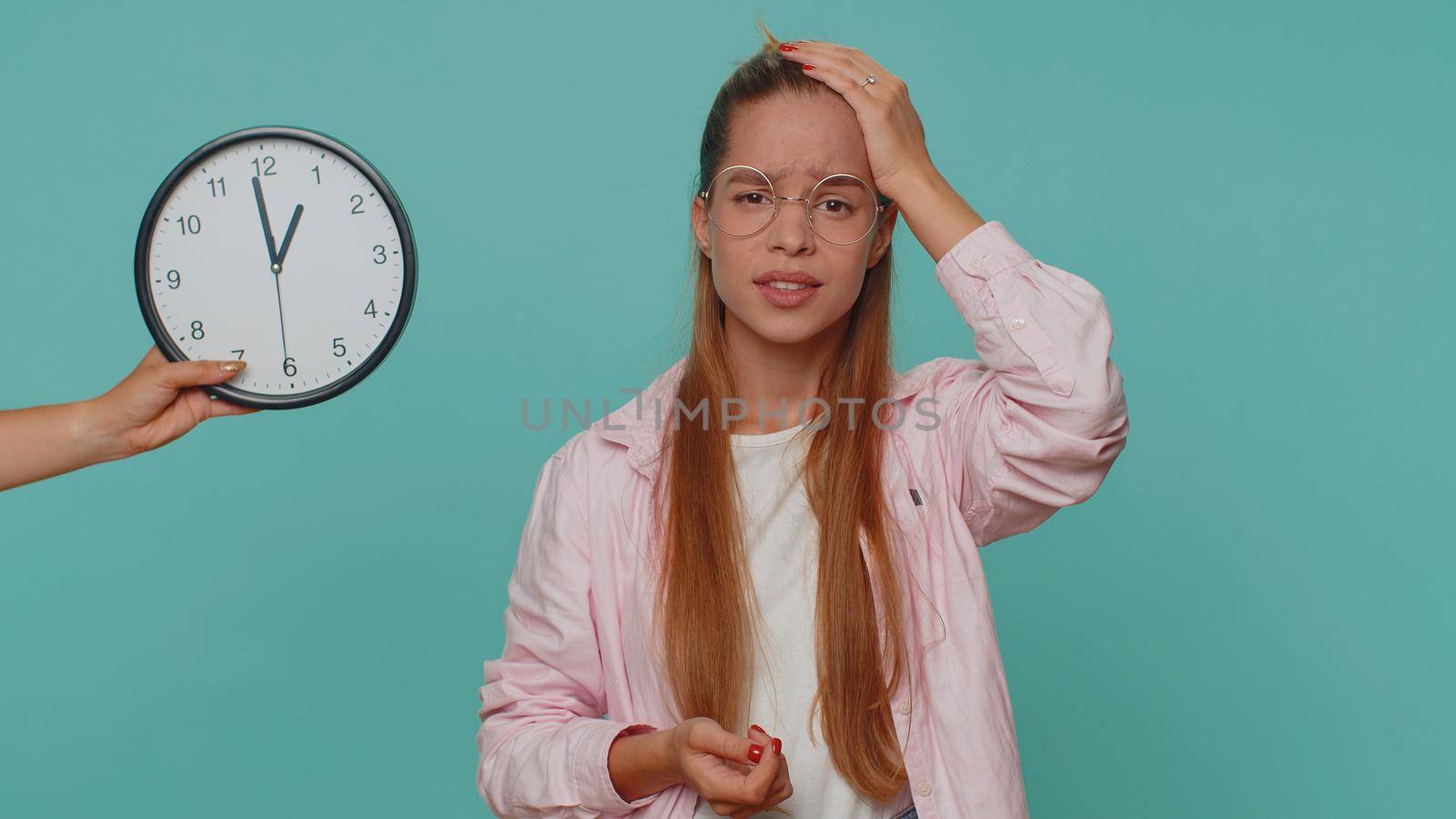 Teenager young girl with anxiety checking time on clock, running late to work, being in delay, deadline. Student child kid looking at hour, minutes, worrying to be punctual on blue studio background