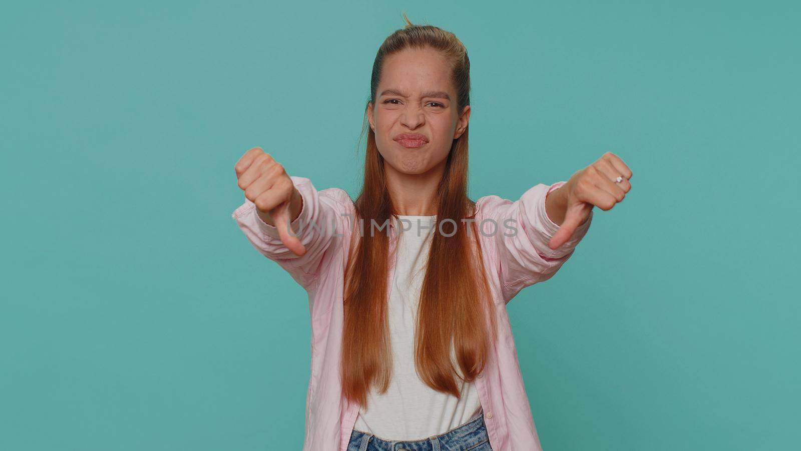 Upset lovely pretty teenager girl showing thumbs down sign gesture, expressing discontent, disapproval, dissatisfied, dislike. Young stylish female child kid isolated alone on blue studio background