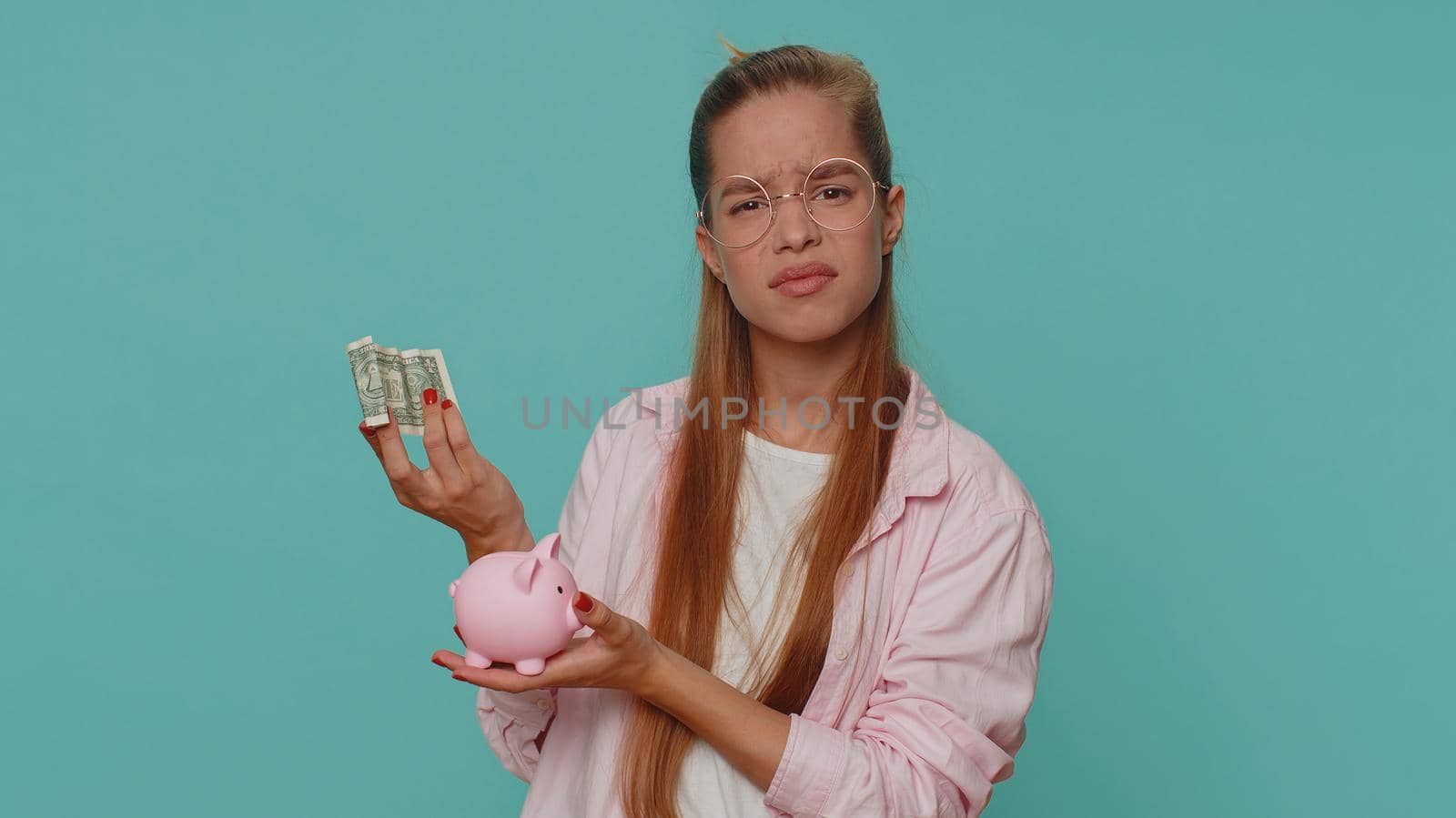 Poor pretty teenager girl insufficient amount of money, holding piggybank and one dollar banknote. Financial crisis. Bankruptcy. Poverty and destitution. Young child kid on blue background indoors
