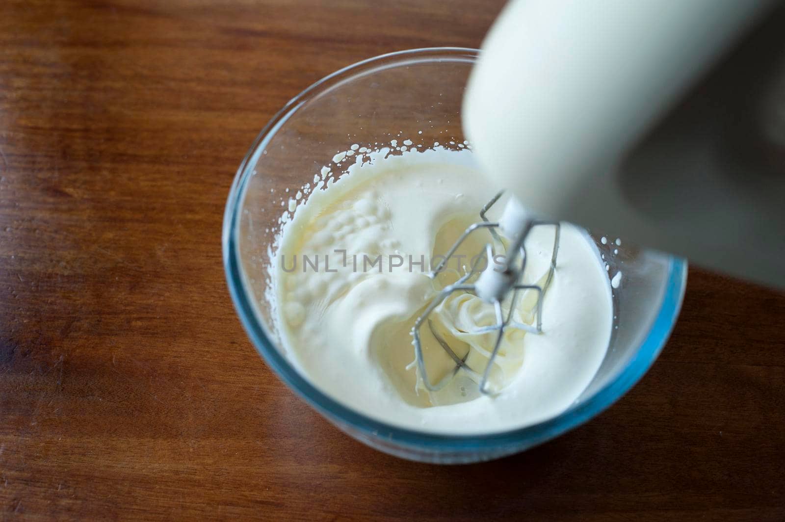 Whipping cream in a glass mixing bowl with a whisk attachment on an electric kitchen mixer, high angle close up view