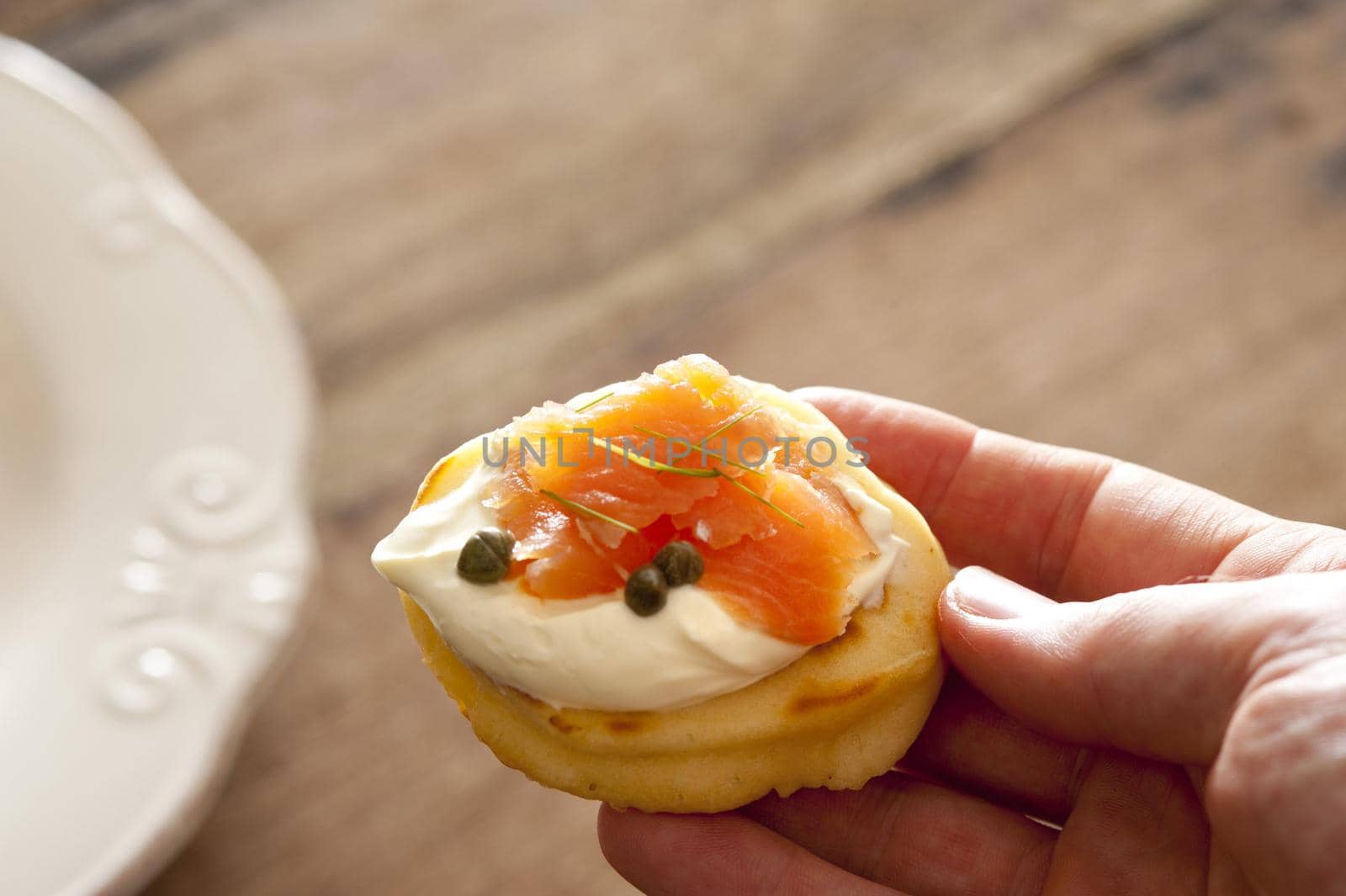 First person view on hand holding delicious blini snack topped with creme and pink salmon over wooden table