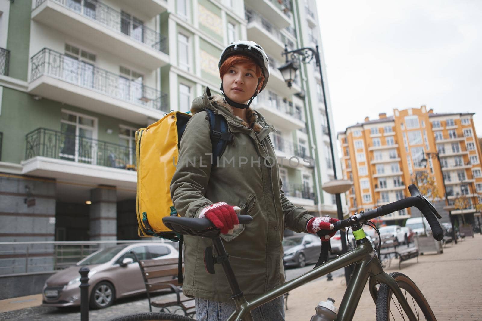 Low angle shot of a female delivery courier walking with her bicycle on city street, wearing delivery backpack