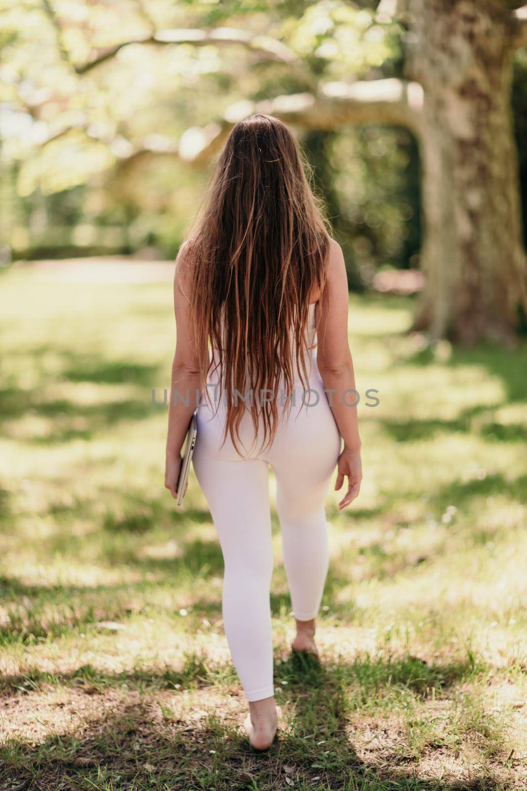 back view of a beautiful woman in a white tracksuit walking in the park with a laptop.