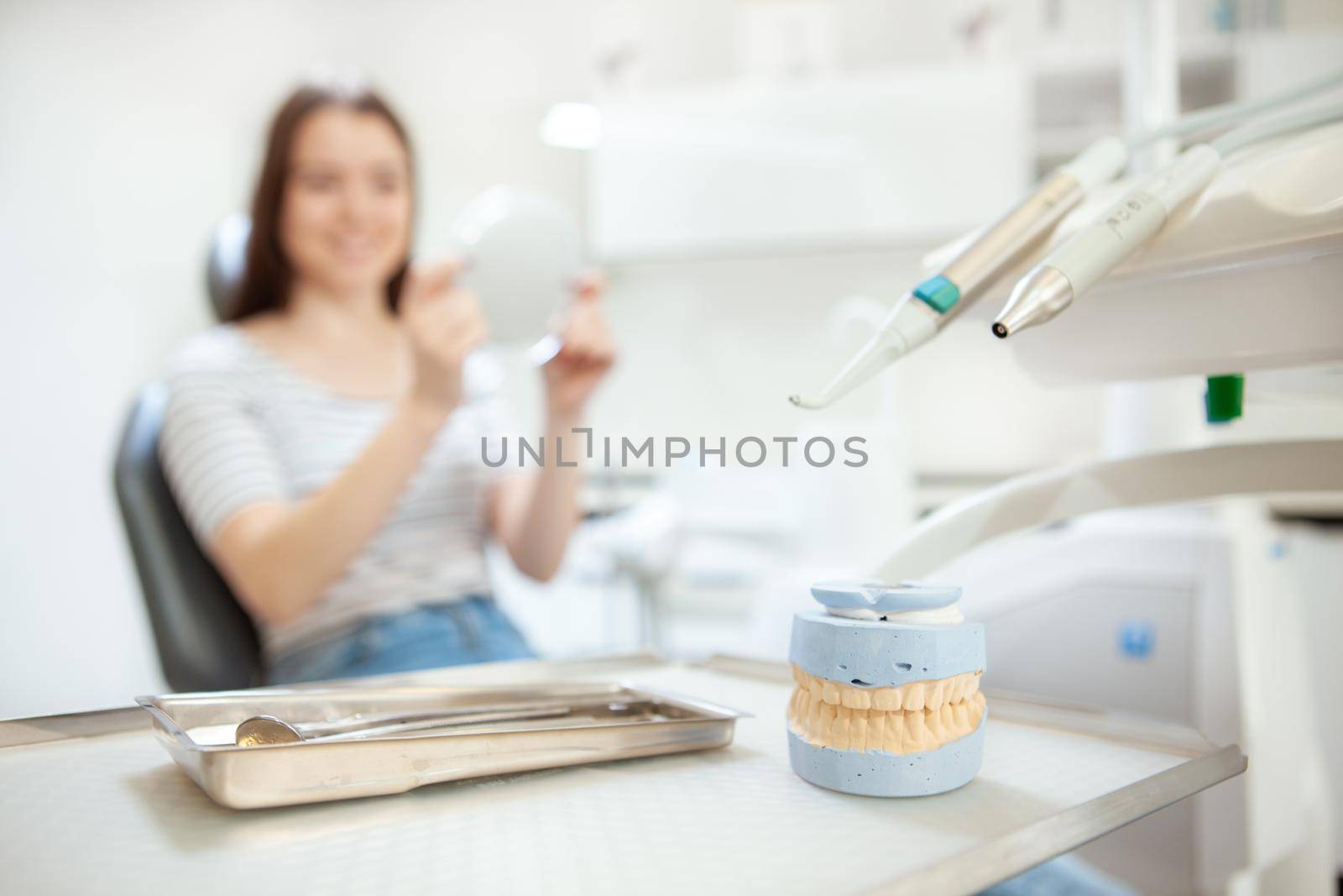 Selective focus on jaw mold and dental equipment, female patient checking her teeth in the mirror after dental treatment on background