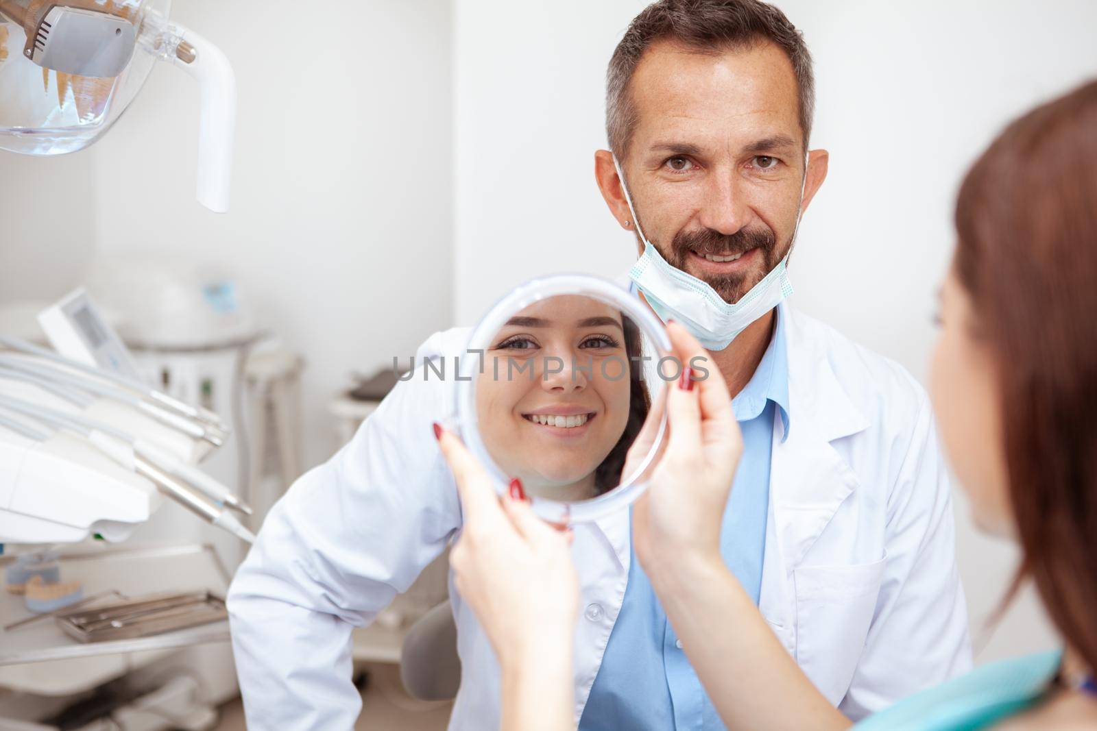 Cheerful mature male dentist smiling to the camera, his female patient checking her teeth in the mirror after dental treatment