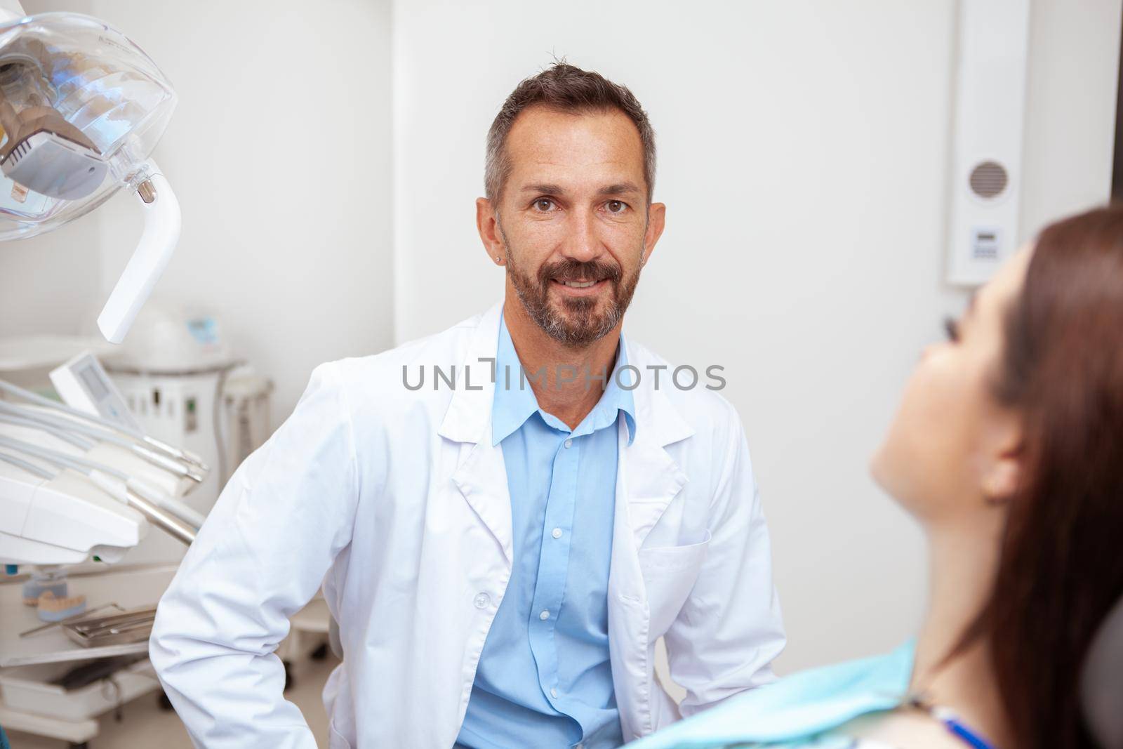 Happy handsome mature male dentist smiling to the camera while working with a patient at his clinic