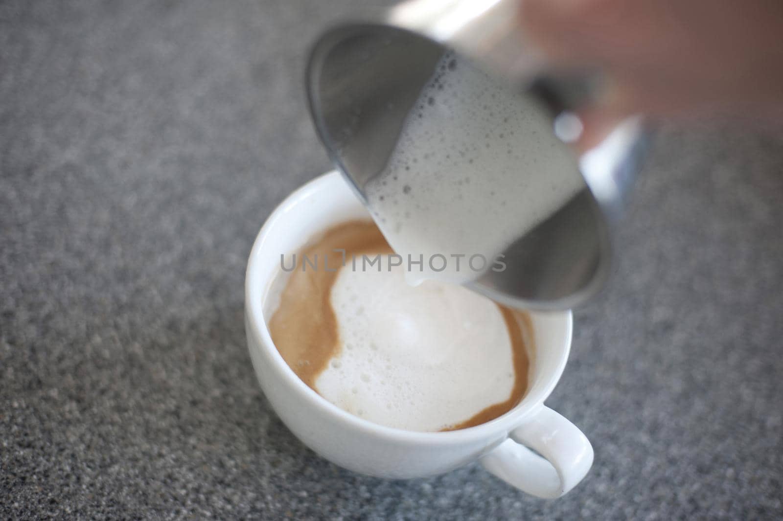 Adding steamed frothy milk to a cup of freshly brewed cappuccino coffee, closeup of a hand pouring the milk from a metal jug, focus to the coffee