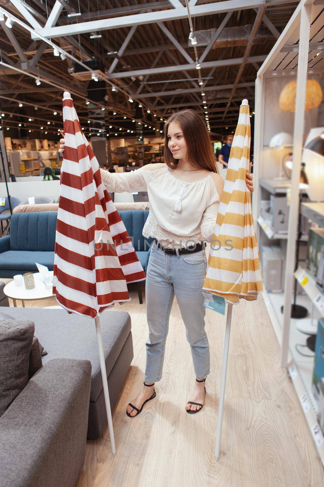 Vertical full length shot of a young woman choosing beach umbrella to buy at furnishings store